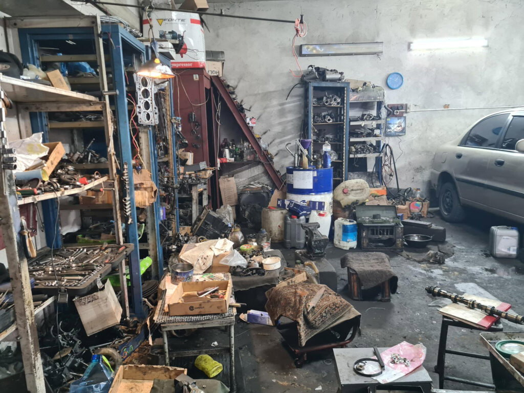 Messy auto repair shop with car parts, tools, shelves, and a silver car partially visible on the right—perfect for a garage cleanout transformation.
