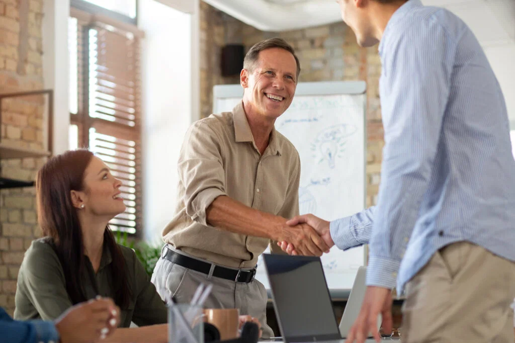 Older man smiling and shaking hands with another person in an office, while a woman sits and watches&mdash;celebrating a successful furniture removal.